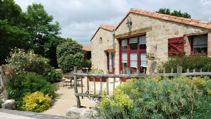 A stone building with red shutters, a wooden fence, and a garden with various plants and flowers. - Bed and Breakfast 'Chambre Nature' with Shared Garden and Wi-Fi (Saint-Malô-du-Bois)