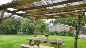 A wooden picnic table with benches under a pergola with a thatched roof, surrounded by a grassy yard with a wooden shed and a fence. - Bed and Breakfast 'Chambre Bohème' with Shared Garden and Wi-Fi (Saint-Malô-du-Bois)