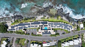 Eine Vogelperspektive zeigt ein Resort mit einer gebogenen Bebauung, einem Swimmingpool und einer Straße, die parallel zur Küstenlinie verläuft.