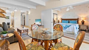 Amplia sala de estar con una mesa de comedor de vidrio, asientos cómodos, una cama grande con ropa de cama azul y un ventilador de techo.
