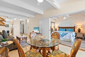 Ein geräumiger Wohnbereich mit einem Glastisch, bequemen Sitzgelegenheiten, einem großen Bett mit blauer Bettwäsche und einem Deckenventilator.