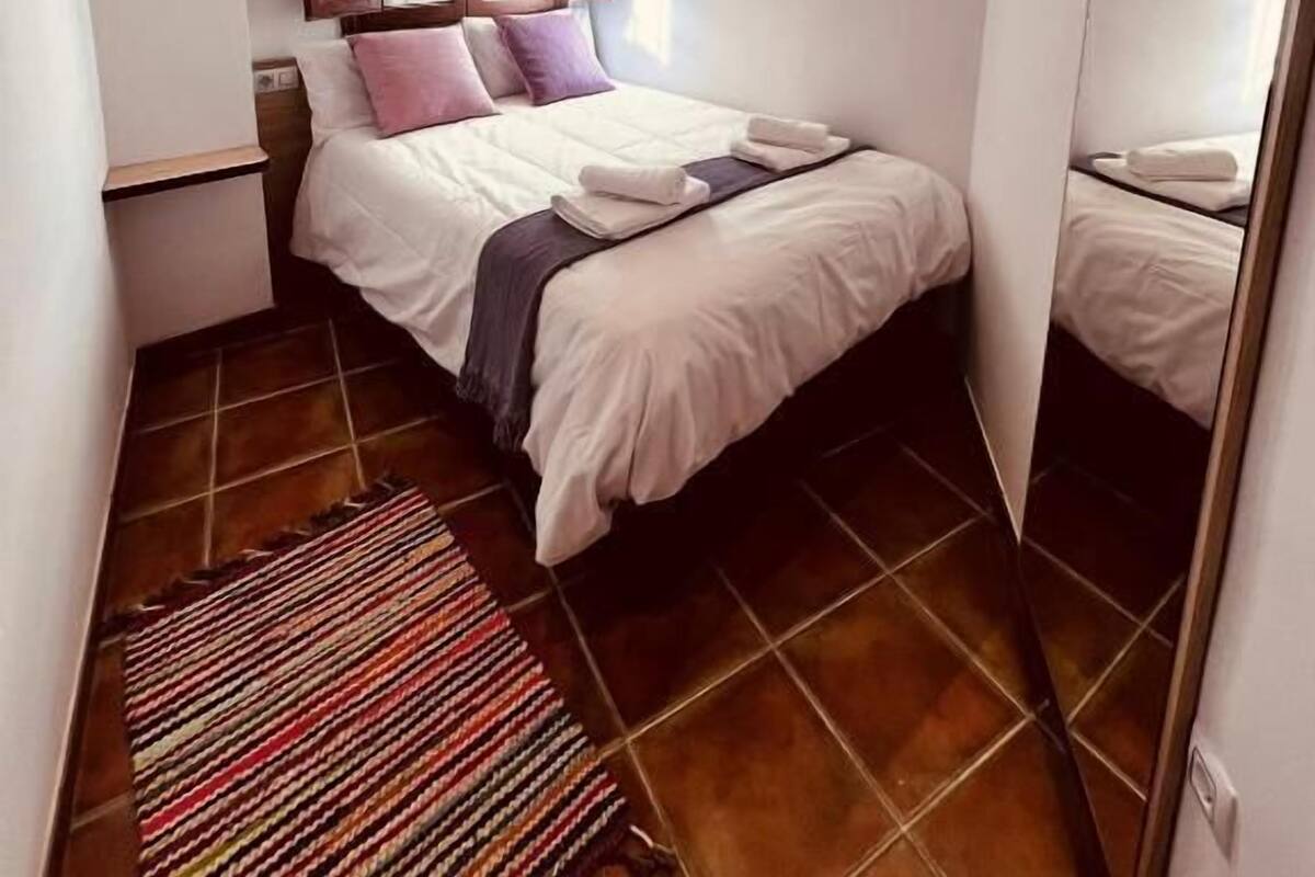 A bedroom with a wooden ceiling, a bed with white bedding, a window with shutters, and a striped rug.