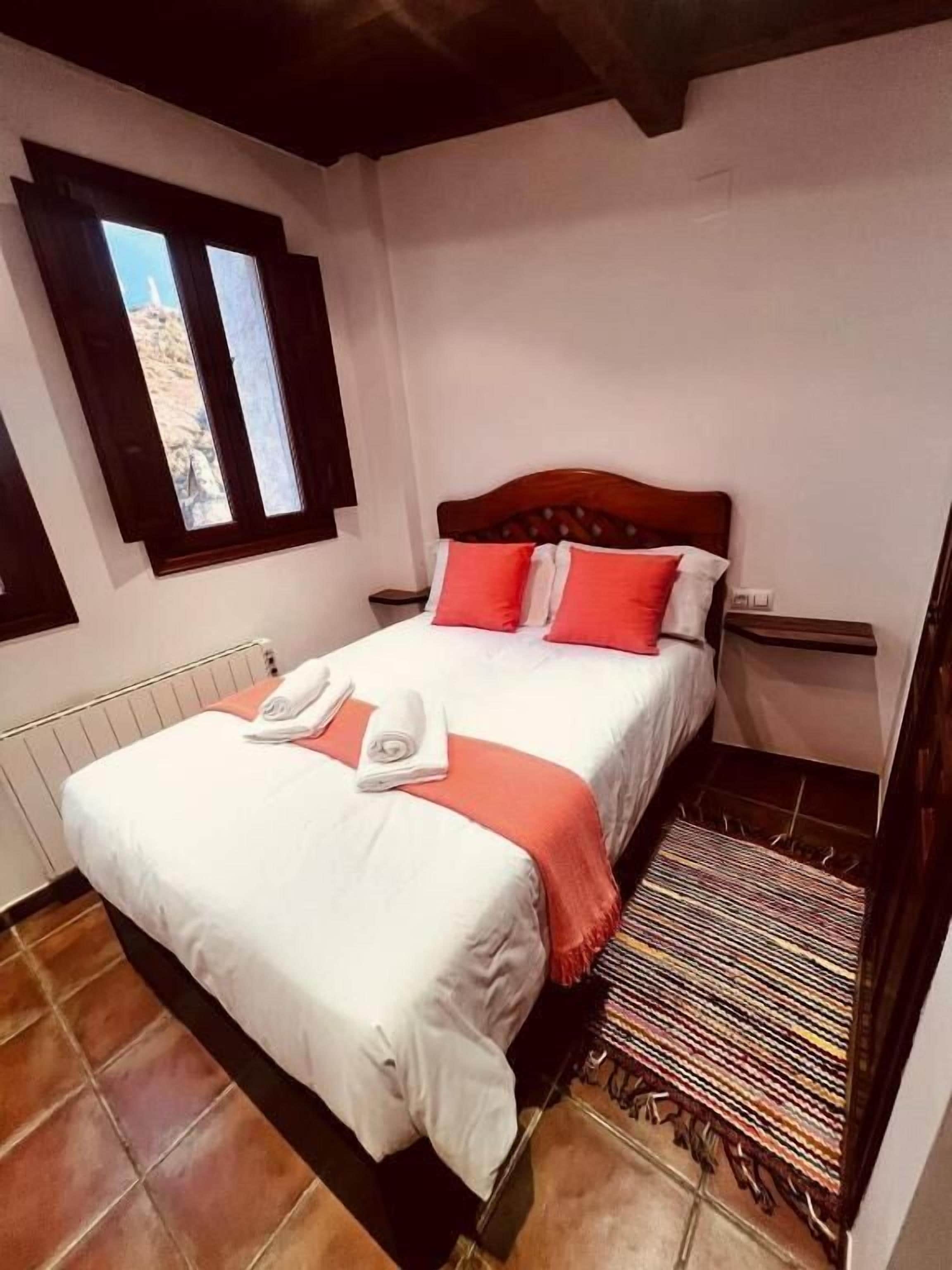 A neatly made bed with white and orange bedding, a wooden headboard, and a window with a view of a rocky landscape.