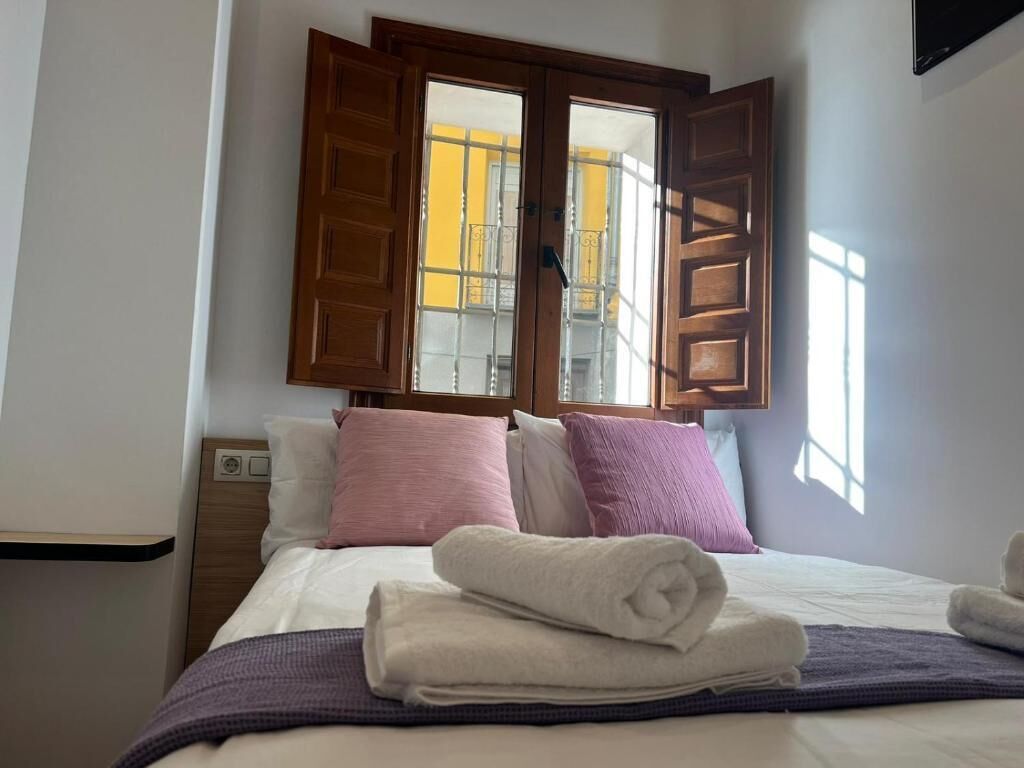 A hotel room with a bed, pillows, a window with wooden shutters, and a view of a building outside.