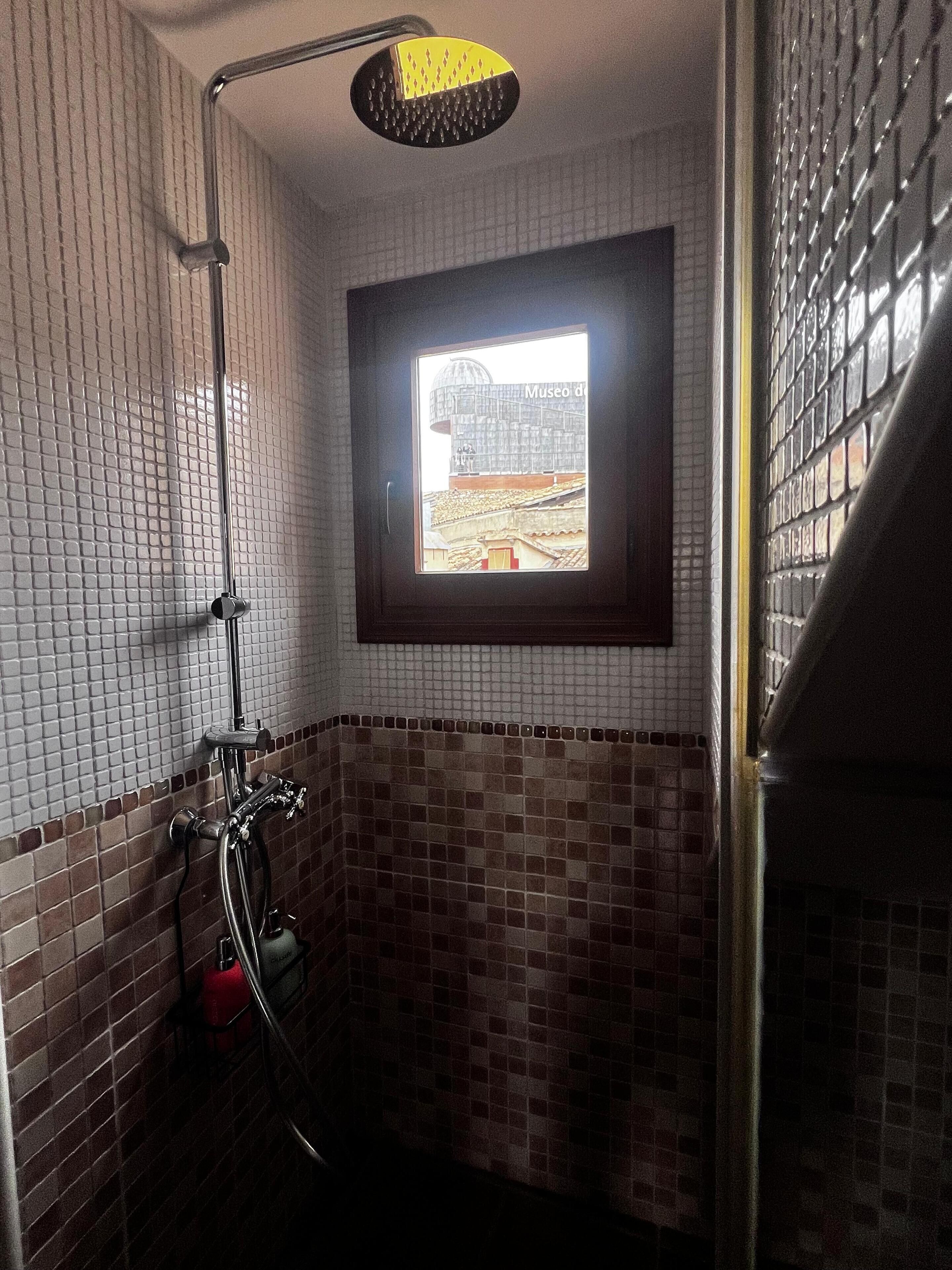 Een badkamer met een douche, een raam met uitzicht op een gebouw en betegelde wanden.