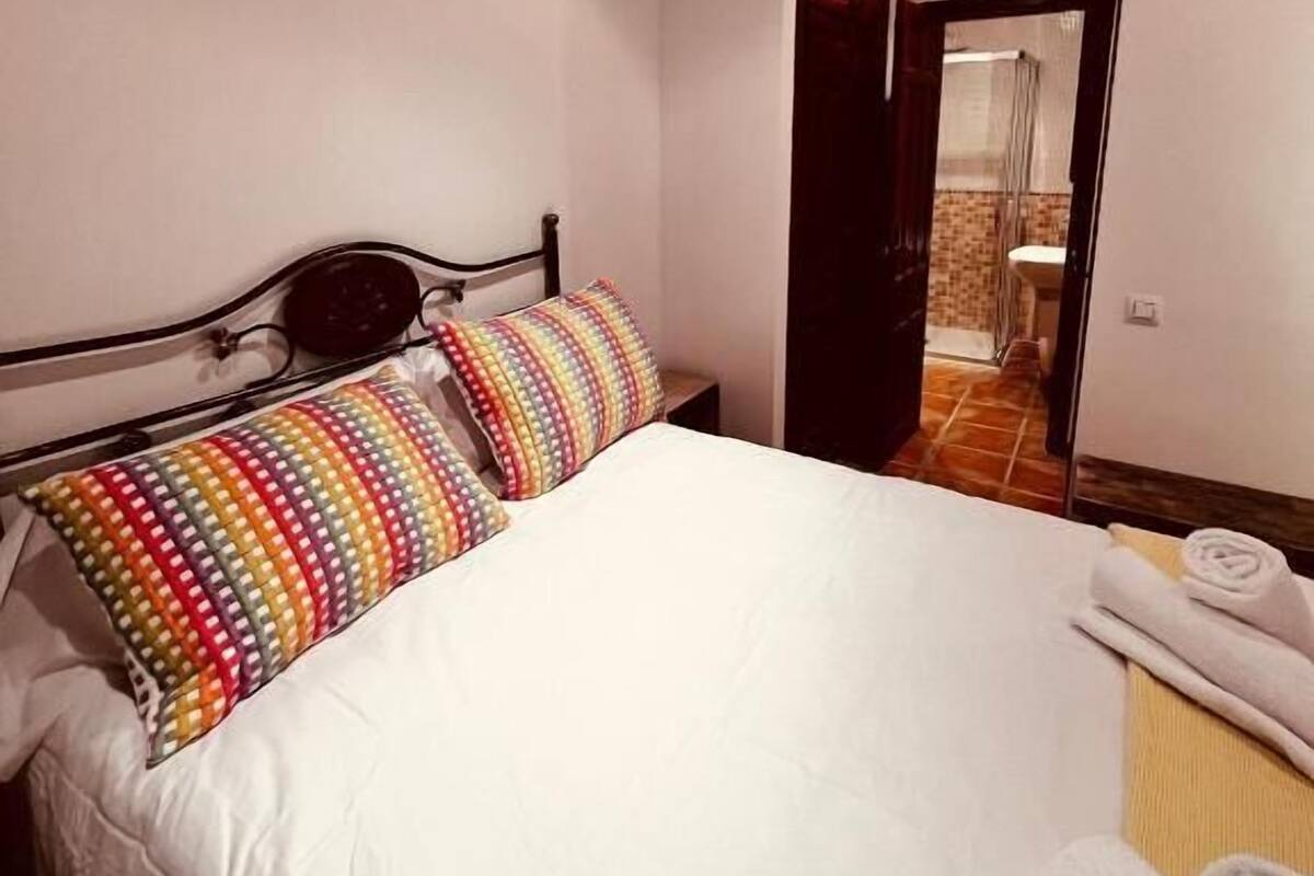 A bedroom with a bed, colorful pillows, a wooden headboard, a white duvet, and a towel on the side.