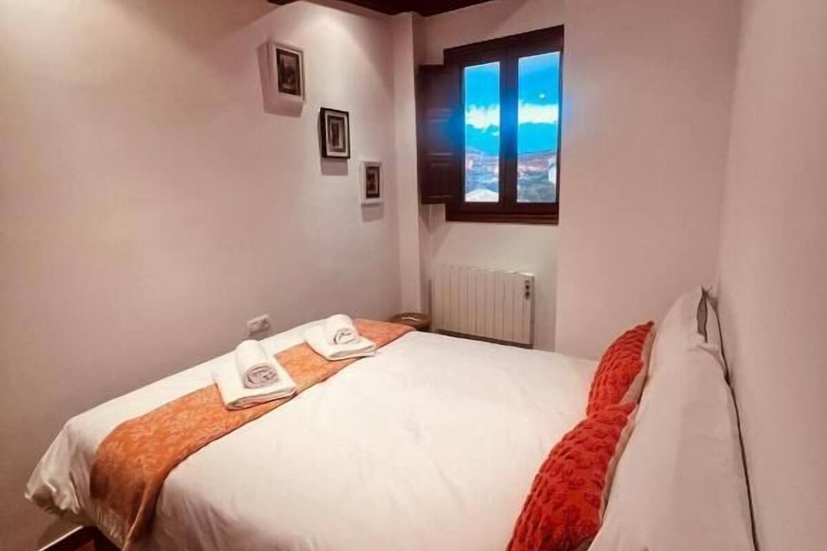 A neatly made bed with white and orange bedding, a window with a view of the sky, and a wooden ceiling.