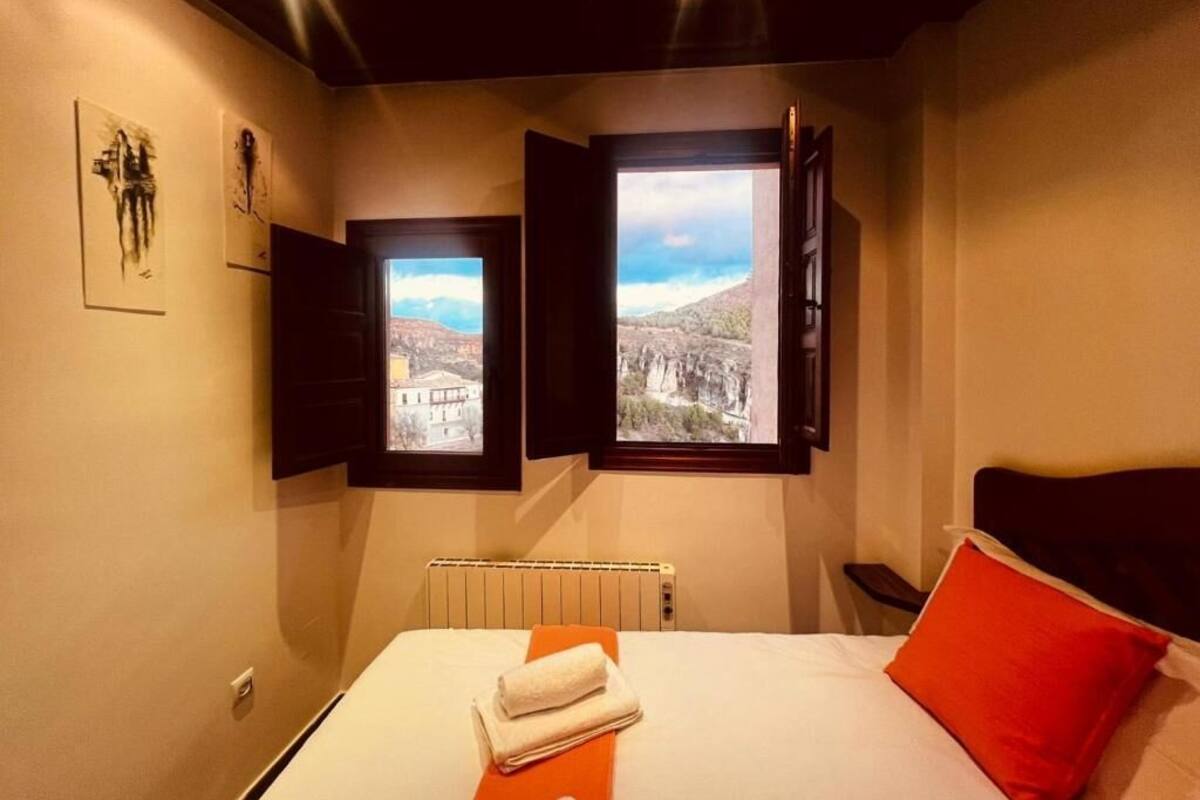 A bedroom with a bed, a red pillow, and two windows with views of a mountainous landscape.