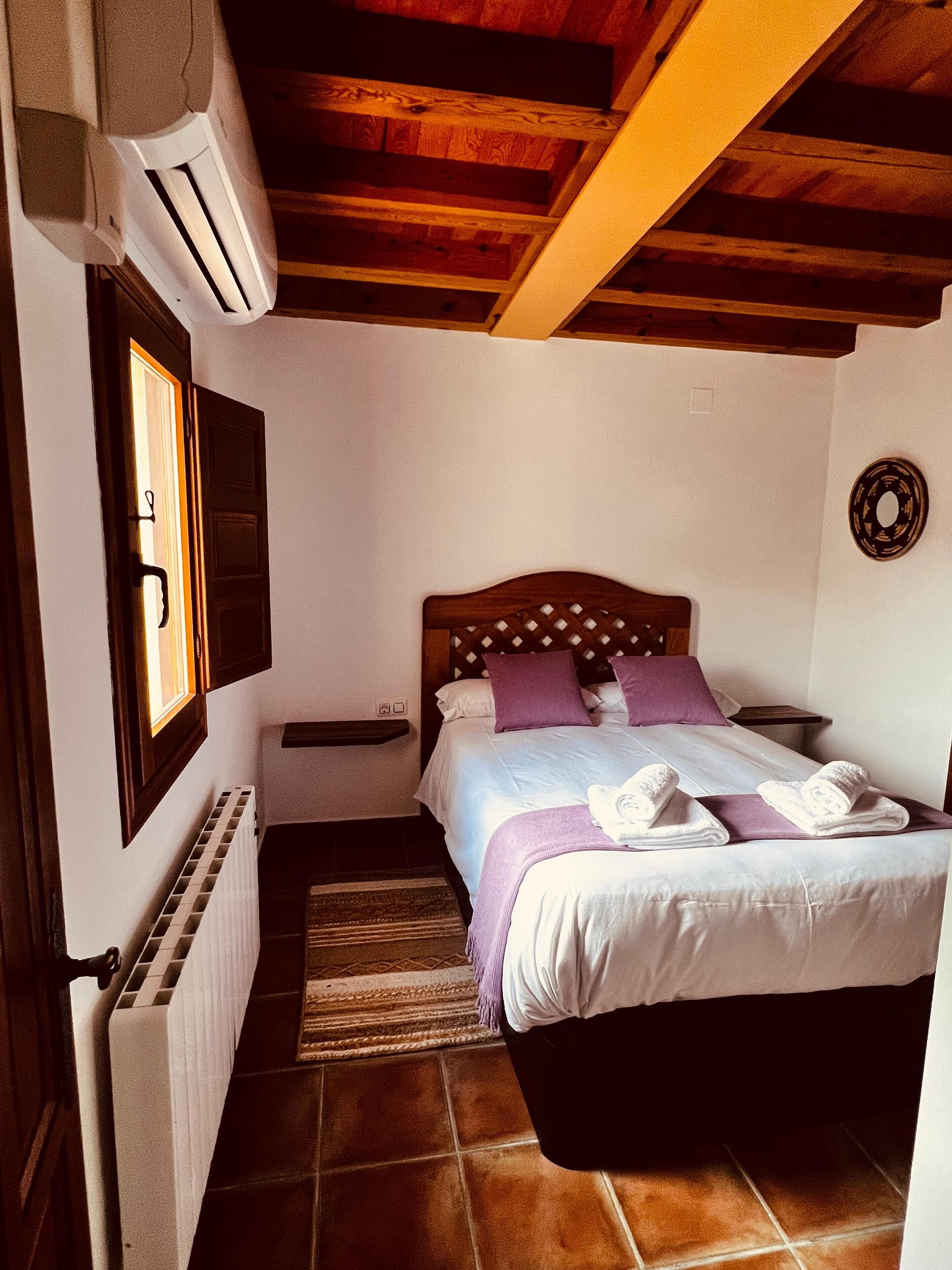 A bedroom with a wooden ceiling, a bed with white and purple bedding, a radiator, and a window with blinds.