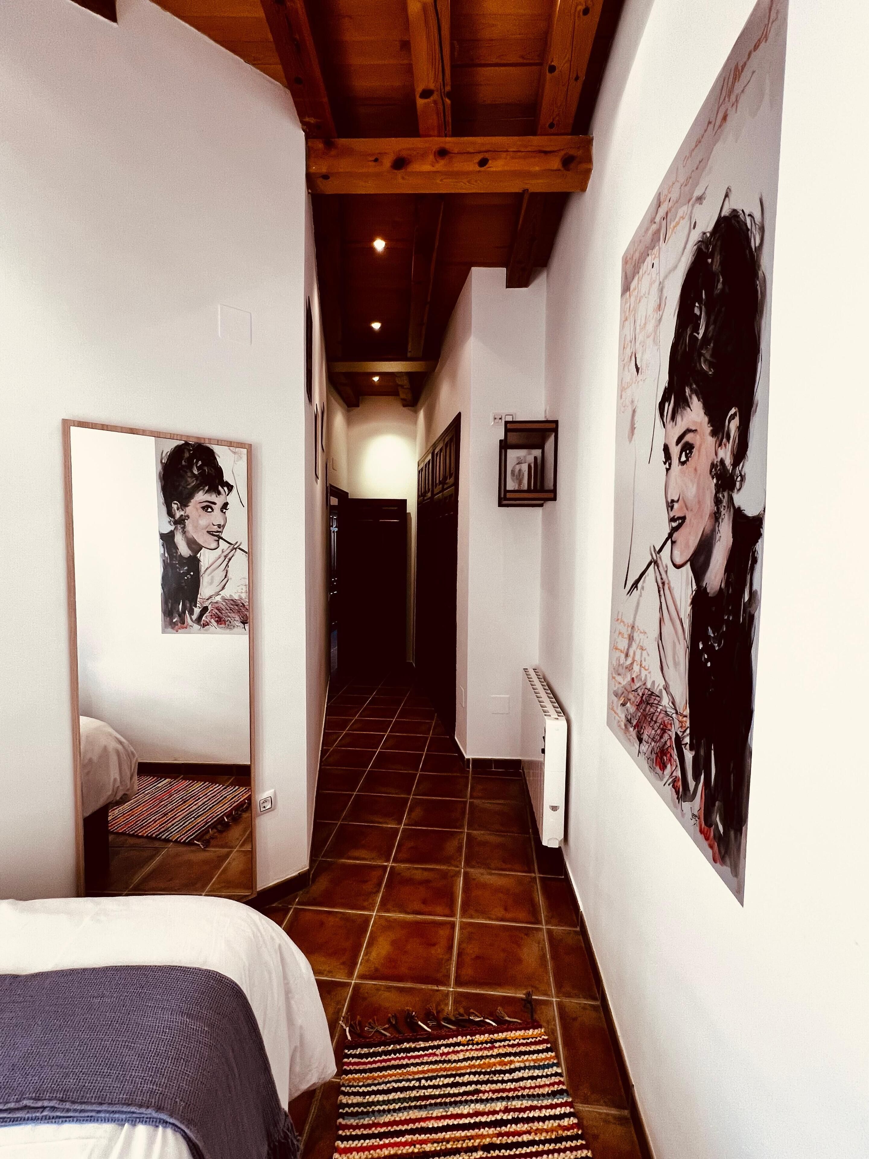 A narrow hallway with a bed on the left, a radiator on the right, and artwork on the walls.
