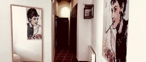 A narrow hallway with a bed on the left, a radiator on the right, and artwork on the walls.