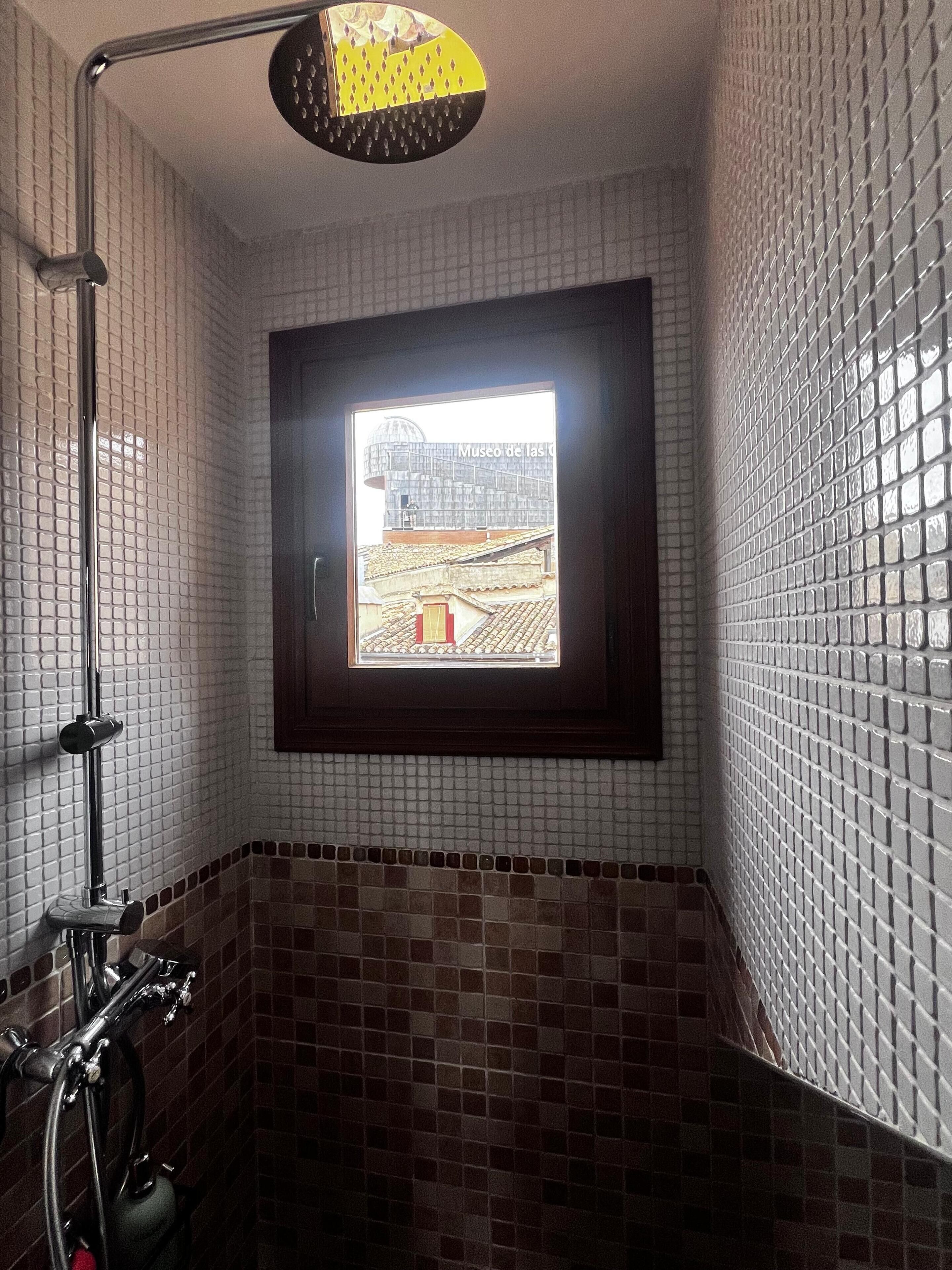 Un baño con ducha, una ventana con vista a un edificio y paredes con azulejos.