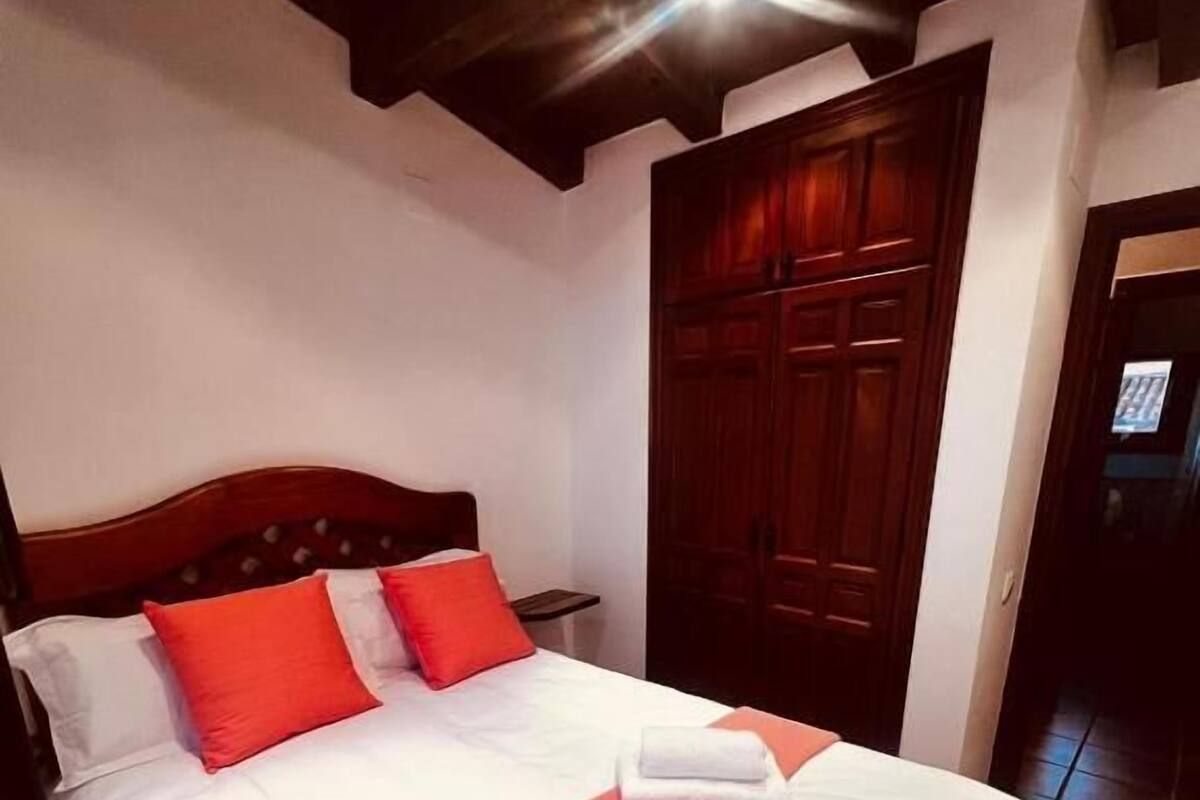 A neatly made bed with white linens and red pillows, a wooden headboard, and a wooden wardrobe in the background.