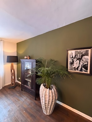 Ein Zimmer mit dunklem Holzboden, einer hohen Stehlampe, einem Schrank mit Glastüren, einer großen dekorativen Pflanzvase und einem gerahmten Schwarz-Weiß-Foto an der Wand.