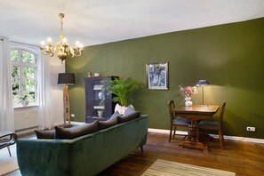 Una sala de estar con un sofá verde, una mesa de comedor con sillas, una araña y un ventanal con cortinas.