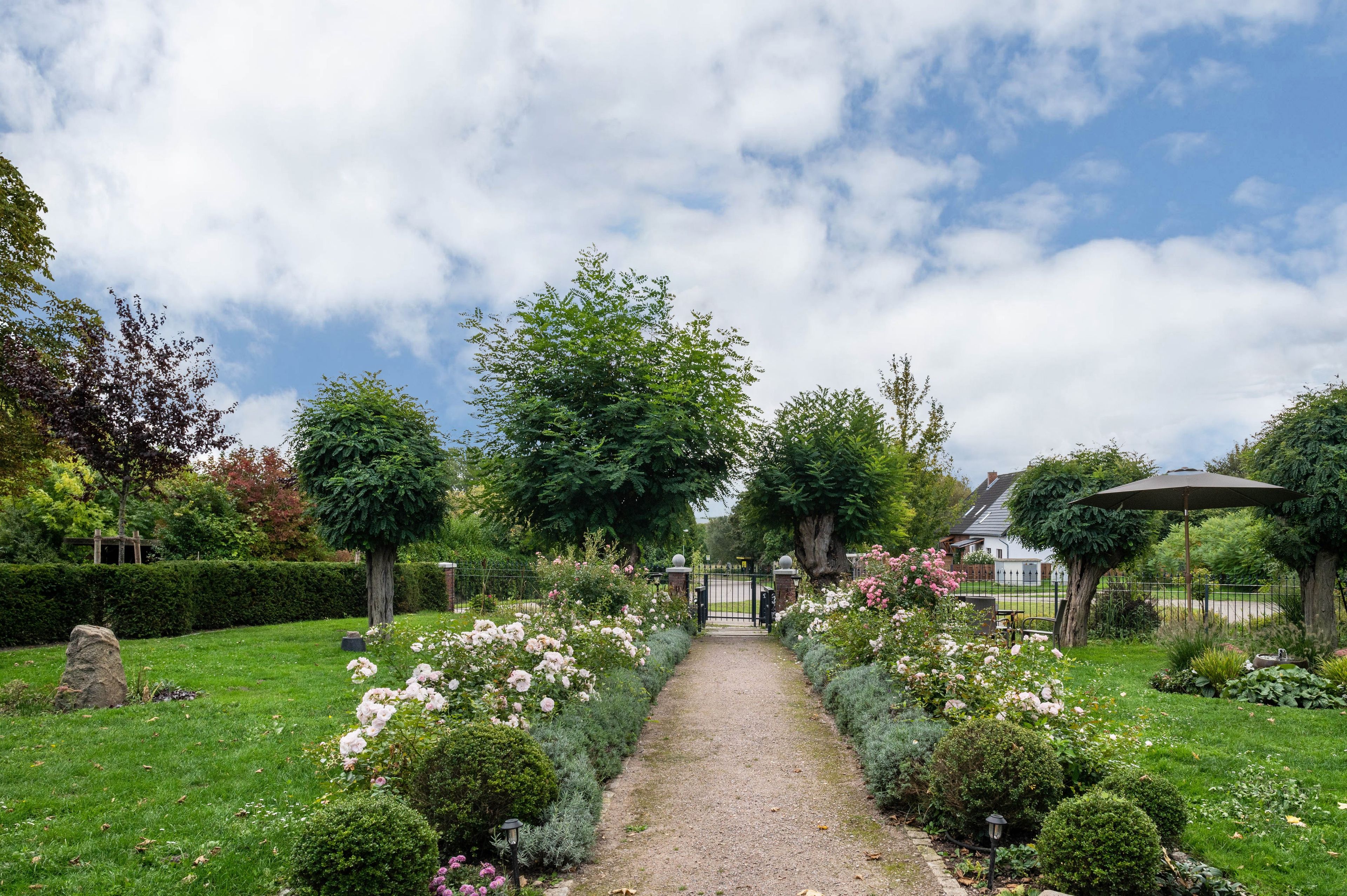 Un sentier de jardin mène à une entrée fermée par un portail, bordée de diverses plantes et arbres en fleurs.