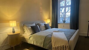 Un dormitorio con cama, lámparas de noche, una ventana con cortinas, un radiador y una alfombra.