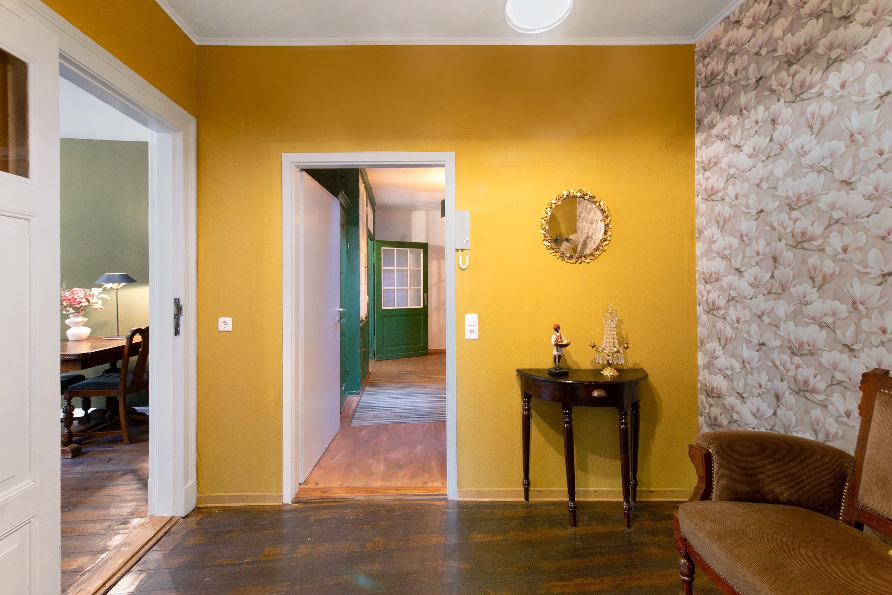 Una habitación con piso de madera, una puerta verde, un espejo y una pequeña mesa con una estatua.
