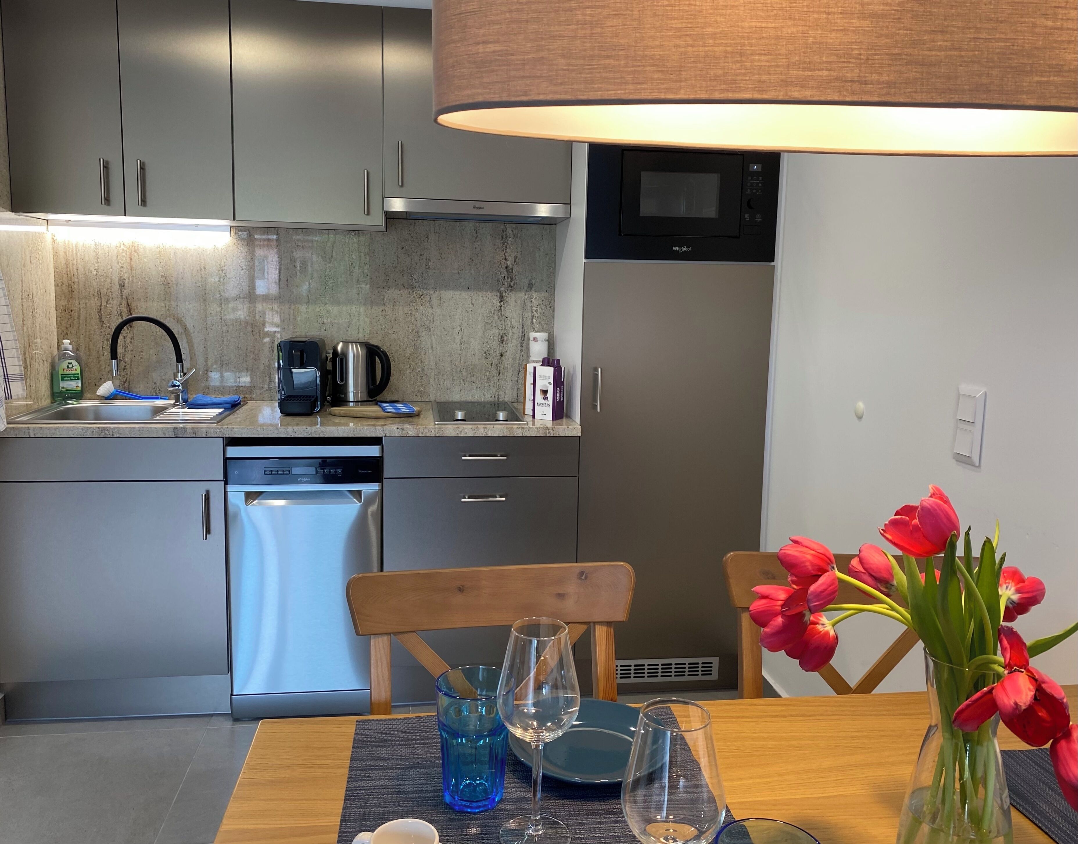 Una cocina moderna con electrodomésticos de acero inoxidable, lavavajillas y microondas. Hay una mesa de comedor con un florero de tulipanes rojos y un mantel individual.