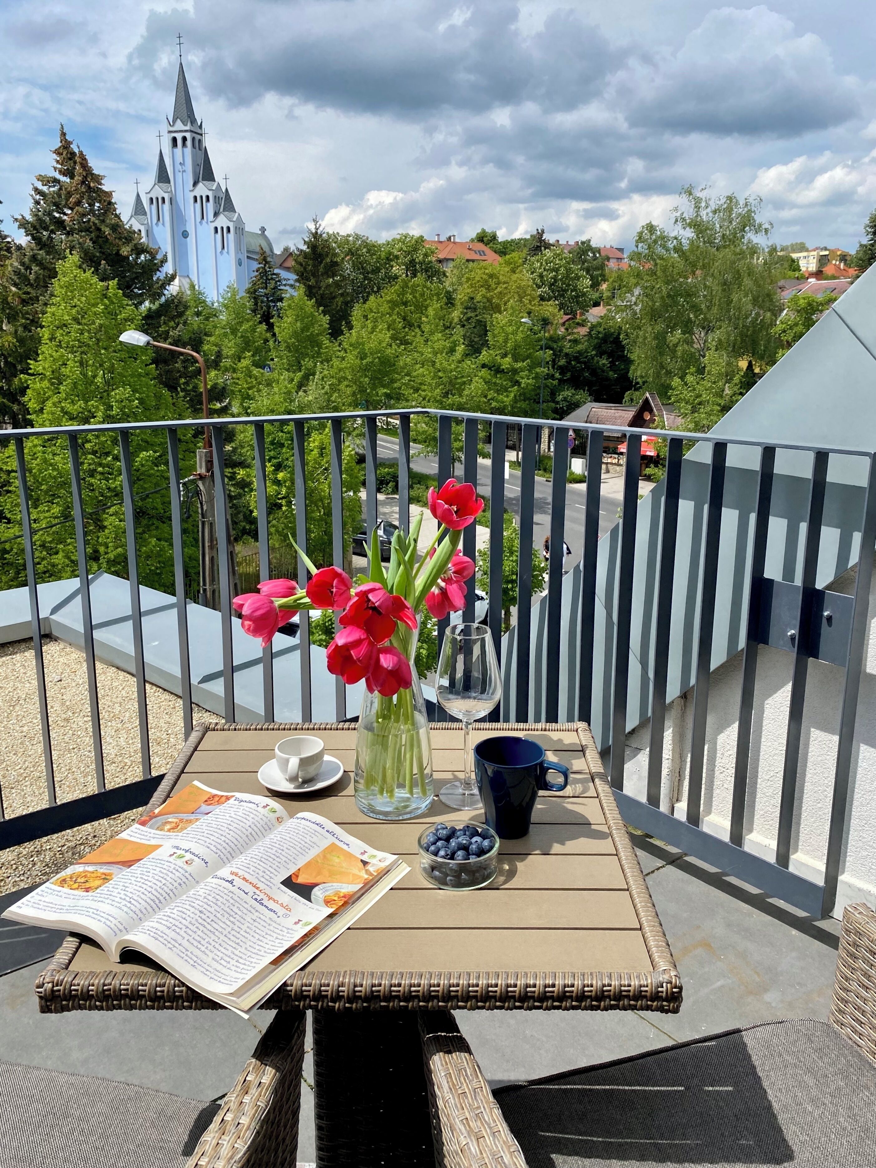 Un balcón con una mesa lista para una comida, un florero con flores y vistas a una iglesia y árboles.