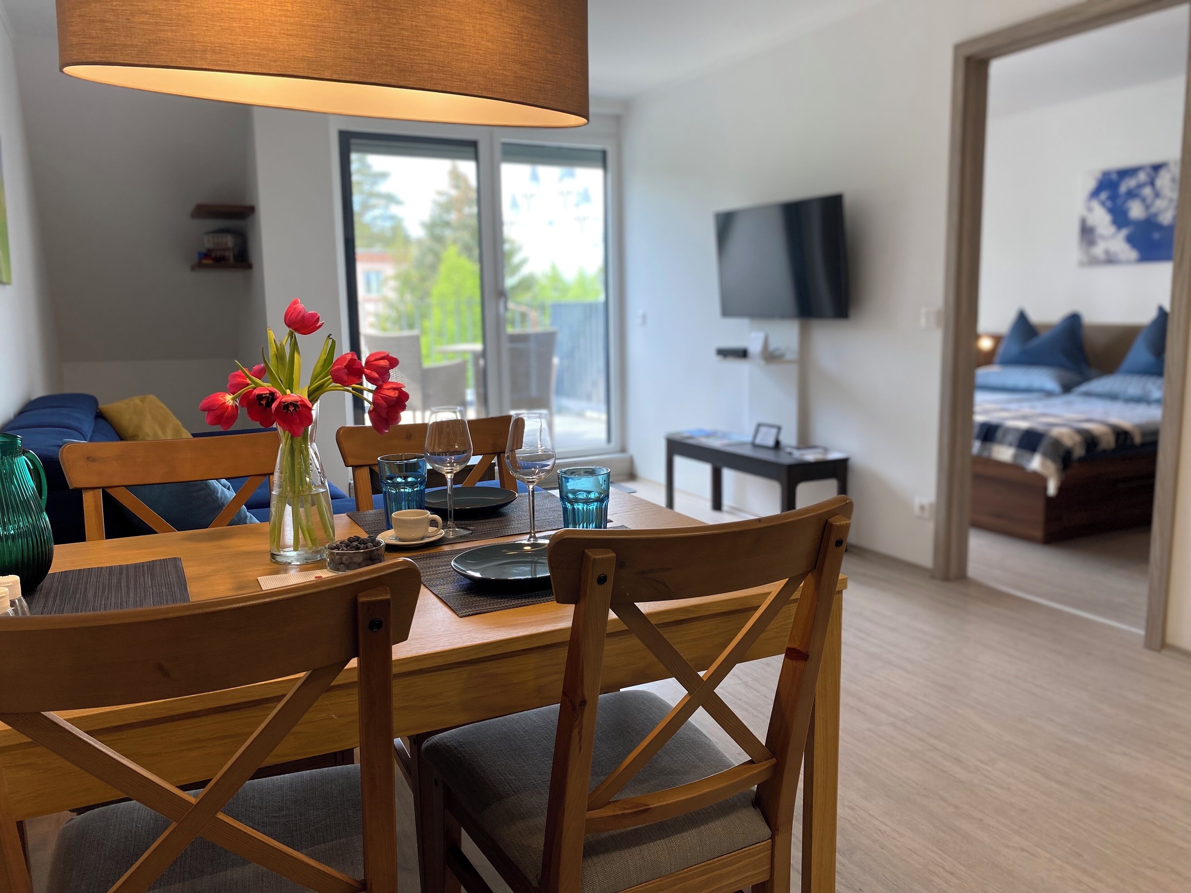 Un coin repas avec une table en bois, des chaises et un vase de tulipes rouges. Un téléviseur est accroché au mur, et on aperçoit un lit avec des oreillers bleus au fond.
