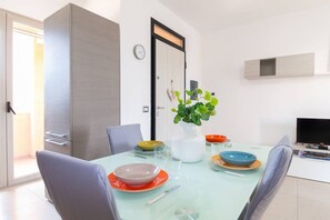 Zona pranzo moderna con tavolo in vetro, sedie, un vaso con foglie verdi e un televisore appeso al muro.