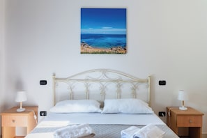A hotel room with a bed, two bedside tables, a nightstand, and a wall-mounted painting of a beach scene.
