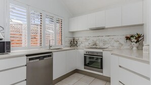 A compact kitchen with white cabinets, a dishwasher, an oven, and a window with blinds.