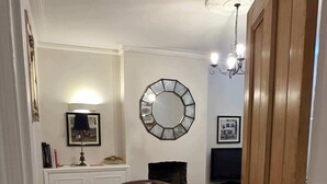 A hallway with wooden flooring, a brown armchair, a mirror, a chandelier, and a television. - Stunning 2-Bed Garden Flat in Fulham (London)