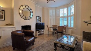 A living room with a brown leather sofa, a glass coffee table, a television, wicker chairs, and a bay window with shutters. - Stunning 2-Bed Garden Flat in Fulham (London)