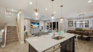 A modern kitchen with a central island, a dining area with a wooden table, and a living room with a sofa and TV. - Beach & Bay by Beach Boutique Rentals (Bradenton Beach)