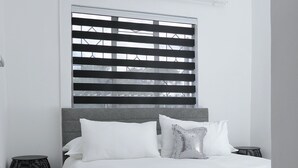 A modern bedroom with a grey headboard, white bedding, a decorative pillow, a bedside table, and a window with blinds. - Motswana Suite: Calm & Cozy 2-bedroom stylish apartment (Gaborone)