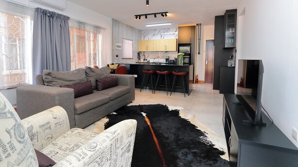 A modern living room with a sofa, a flat-screen TV, and a kitchen area in the background. - Motswana Suite: Calm & Cozy 2-bedroom stylish apartment (Gaborone)