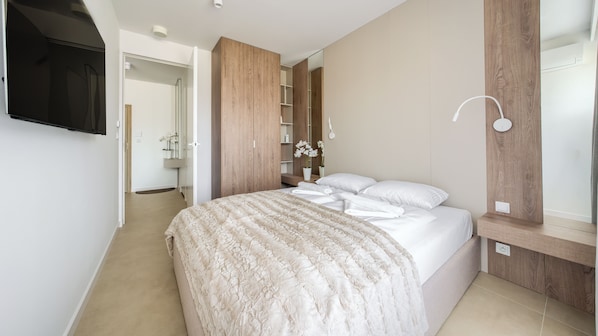 A bedroom with a bed, a TV mounted on the wall, a wooden wardrobe, and a bedside table with a lamp. - Sun & Snow Greeneri Park (Świnoujście)