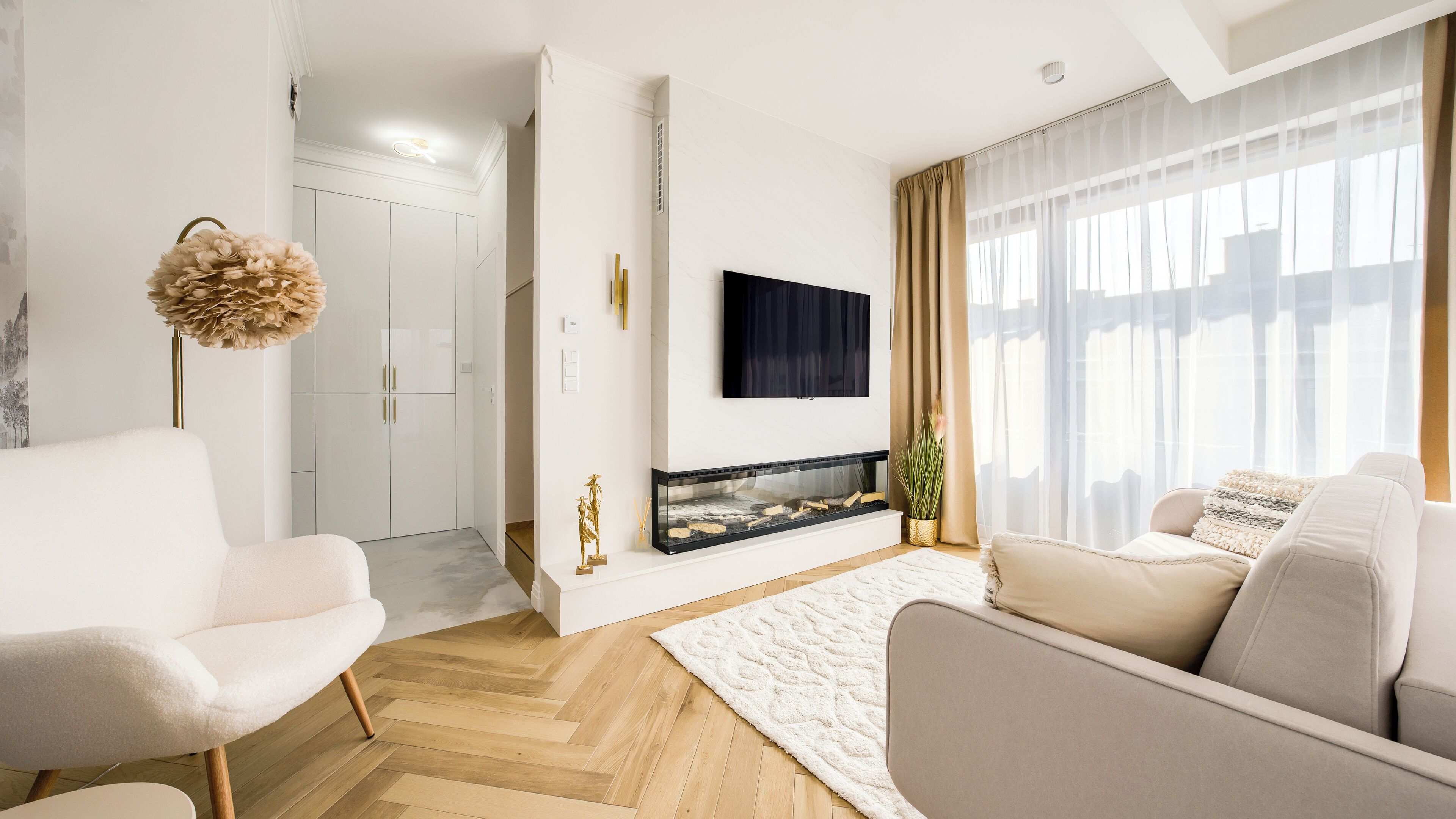 A modern living room with a fireplace, a flat-screen TV mounted on the wall, and a large window with sheer curtains.