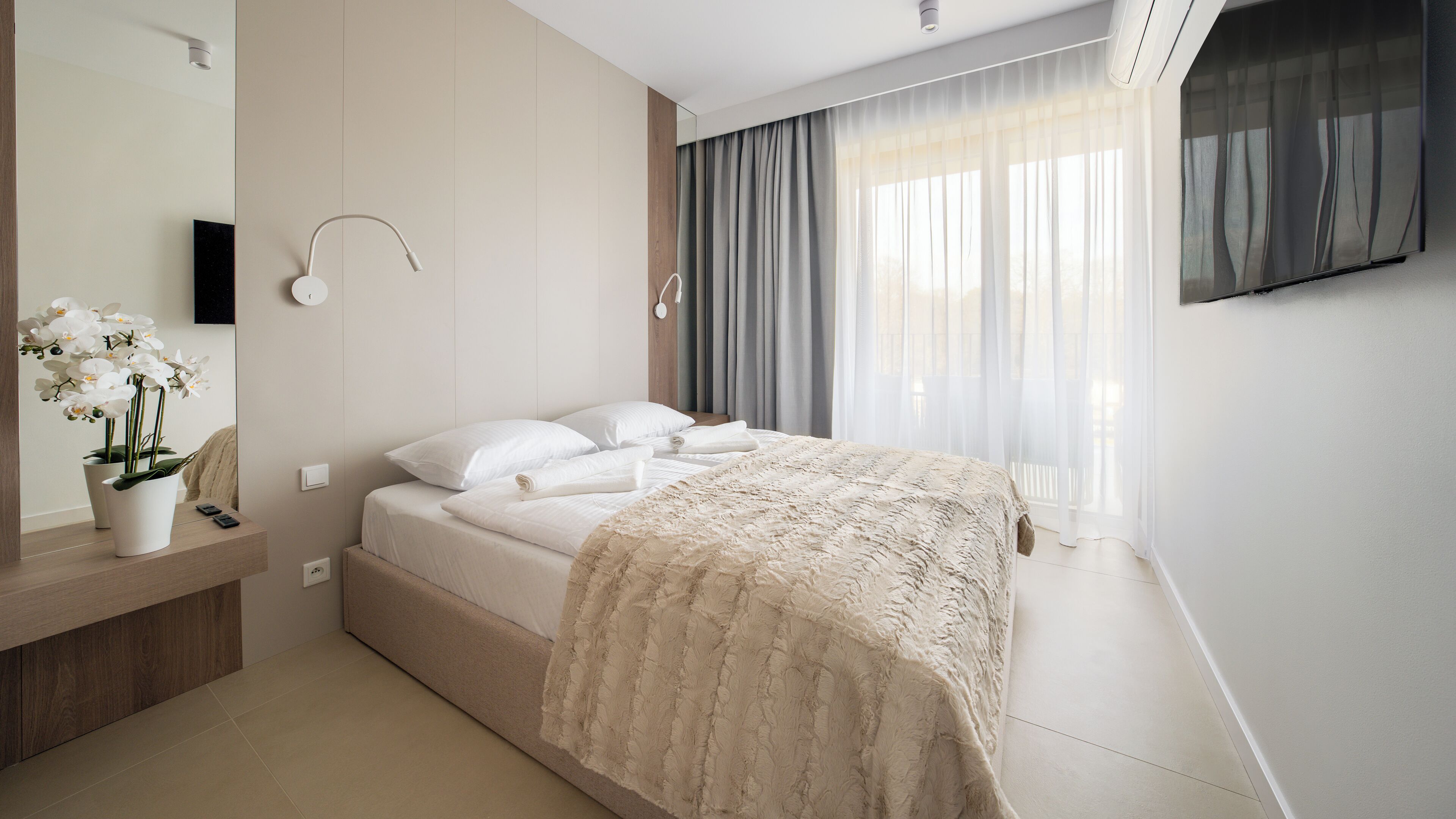 A modern hotel room with a bed, a bedside table with a phone, a mirror, and a potted orchid.