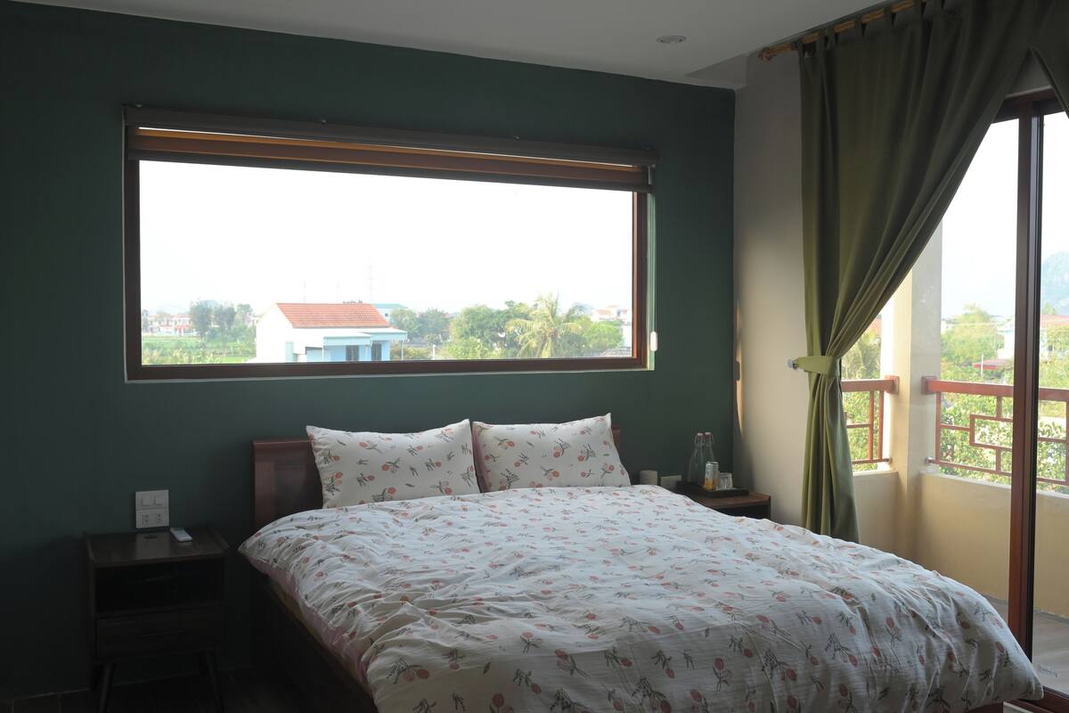 Un dormitorio con un ventanal grande, una cama con ropa de cama floral, una mesita de noche con un teléfono, y vistas a zonas verdes y edificios al exterior.