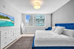 Un dormitorio con cama, televisor, ventana y vistas a una playa y edificios.