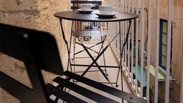 Un étroit couloir extérieur avec une table et des chaises en métal, un banc et une rambarde blanche.