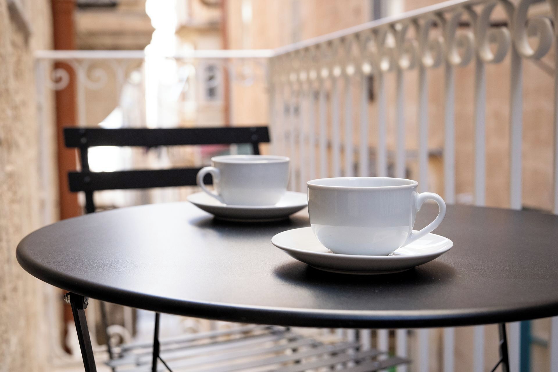 A round table with two white cups and saucers, a black chair, and a metal railing in the background.