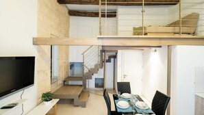 Un appartement moderne de style « loft » comprenant un coin repas, un téléviseur et un escalier menant à une mezzanine aménagée en espace nuit.