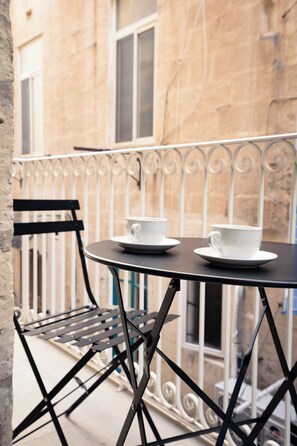 Un balcon avec une table ronde et deux tasses blanches posées sur des soucoupes, une chaise en métal noir et une balustrade en fer forgé.