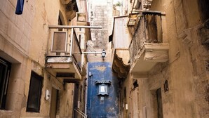 Une ruelle étroite pavée de briques, bordée de bâtiments dotés de balcons, et au loin, un mur bleu.