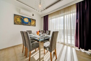 Un coin repas comprenant une table en verre, quatre chaises grises à revêtement rembourré, un lustre et un grand tableau abstrait accroché au mur.