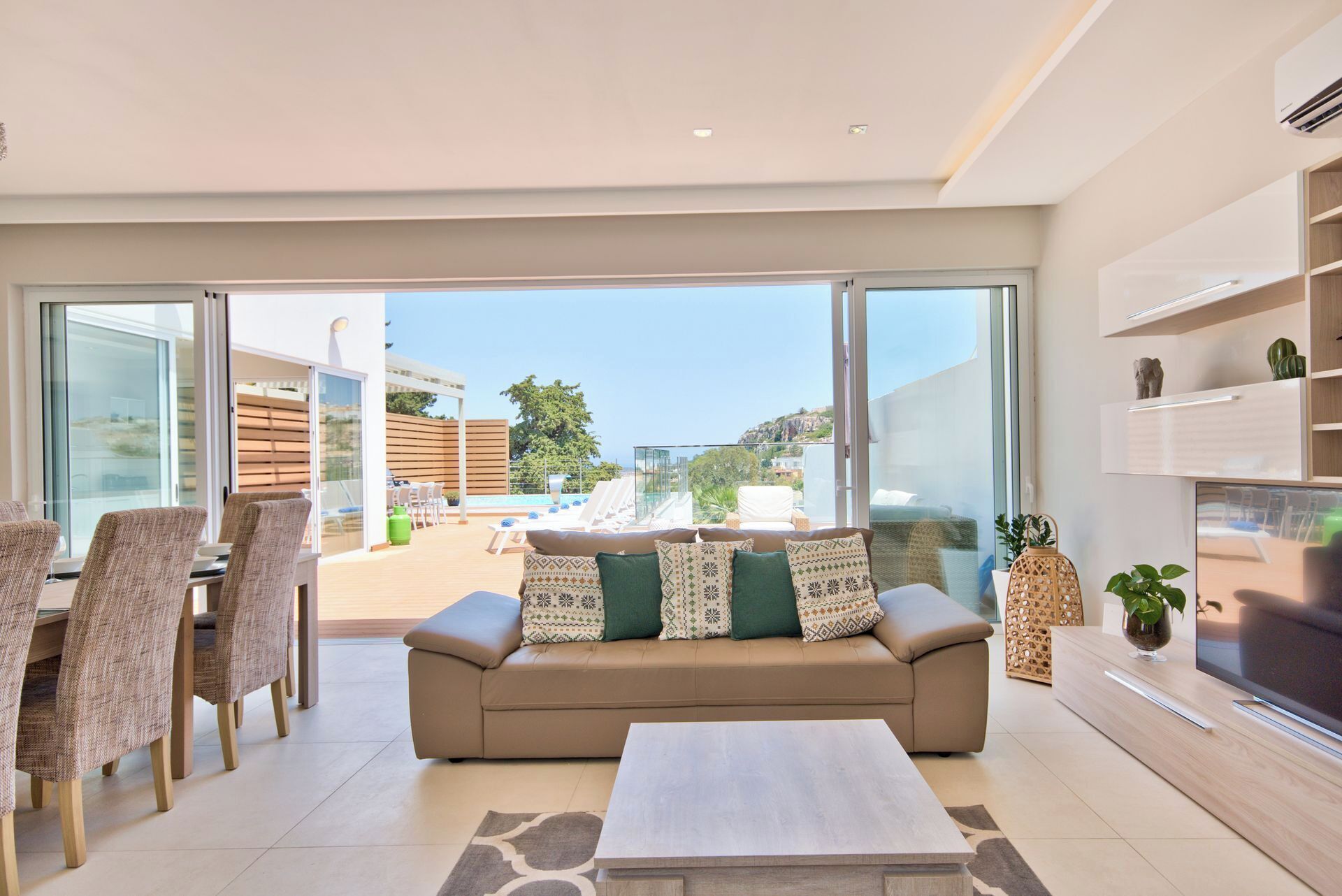 A modern living room with a sofa, chairs, and a TV. There is a balcony with a pool and lounge chairs.