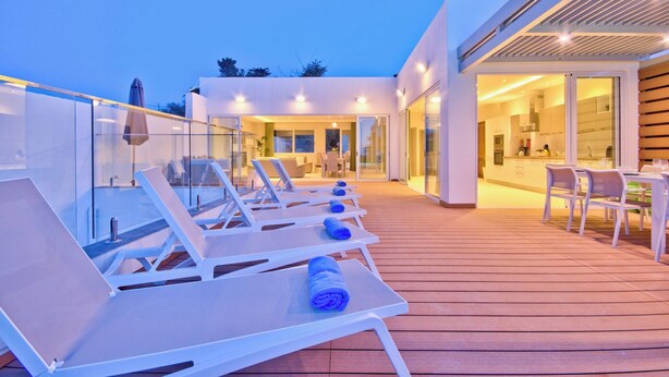 A modern outdoor deck with sun loungers and a glass balustrade, leading to a contemporary interior with a kitchen and dining area. - Extraordinary Villa With Private Pool & Steamsauna (Mellieha)