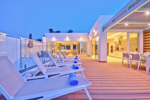A modern outdoor deck with sun loungers and a glass balustrade, leading to a contemporary interior with a kitchen and dining area. - Extraordinary Villa With Private Pool & Steamsauna (Mellieha)