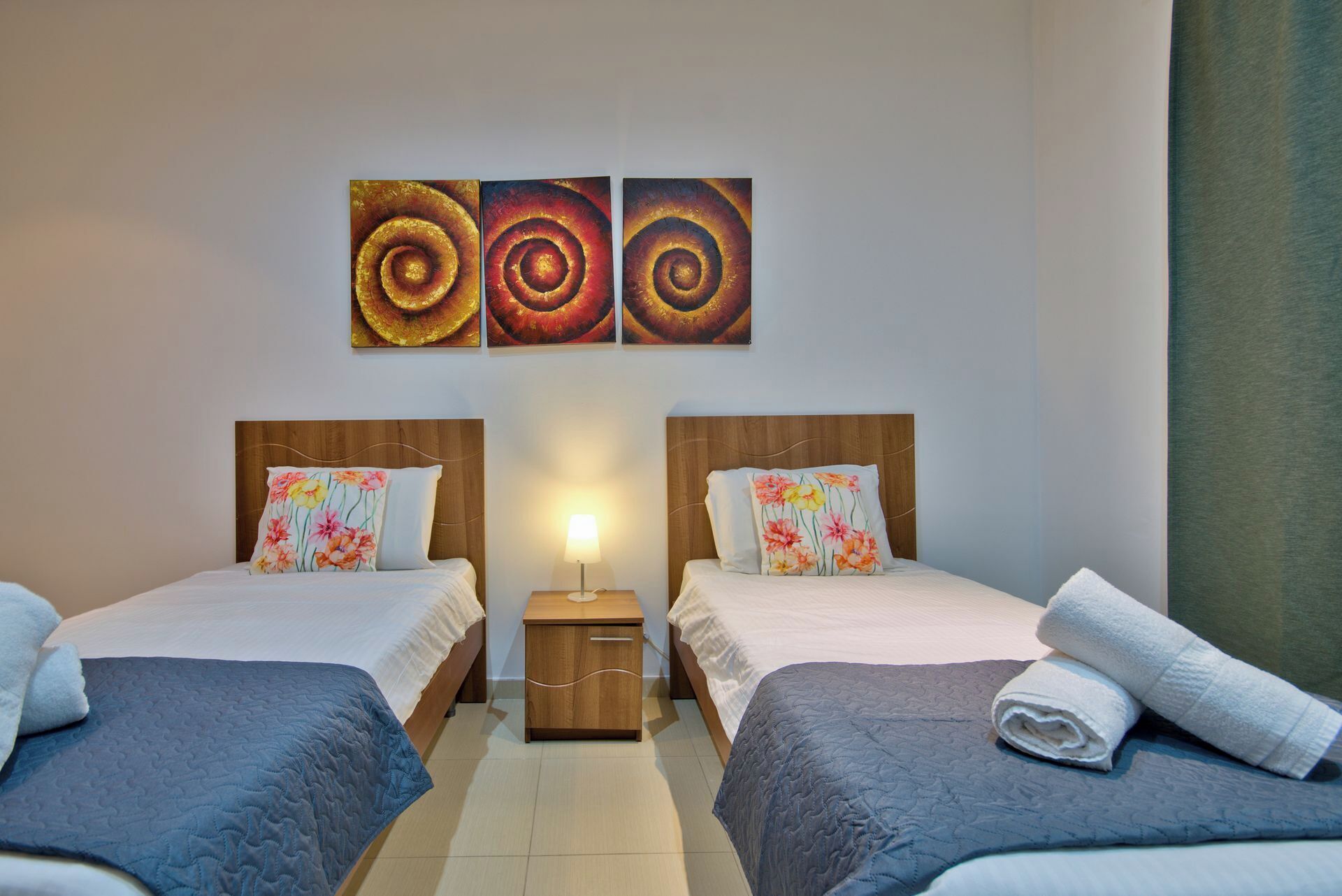 Ein Hotelzimmer mit zwei Betten, jedes mit einem floralen Kopfkissen und einer blauen Steudecke. Darüber hängen drei Wandbilder, dazwischen ein Nachttisch mit einer Lampe.