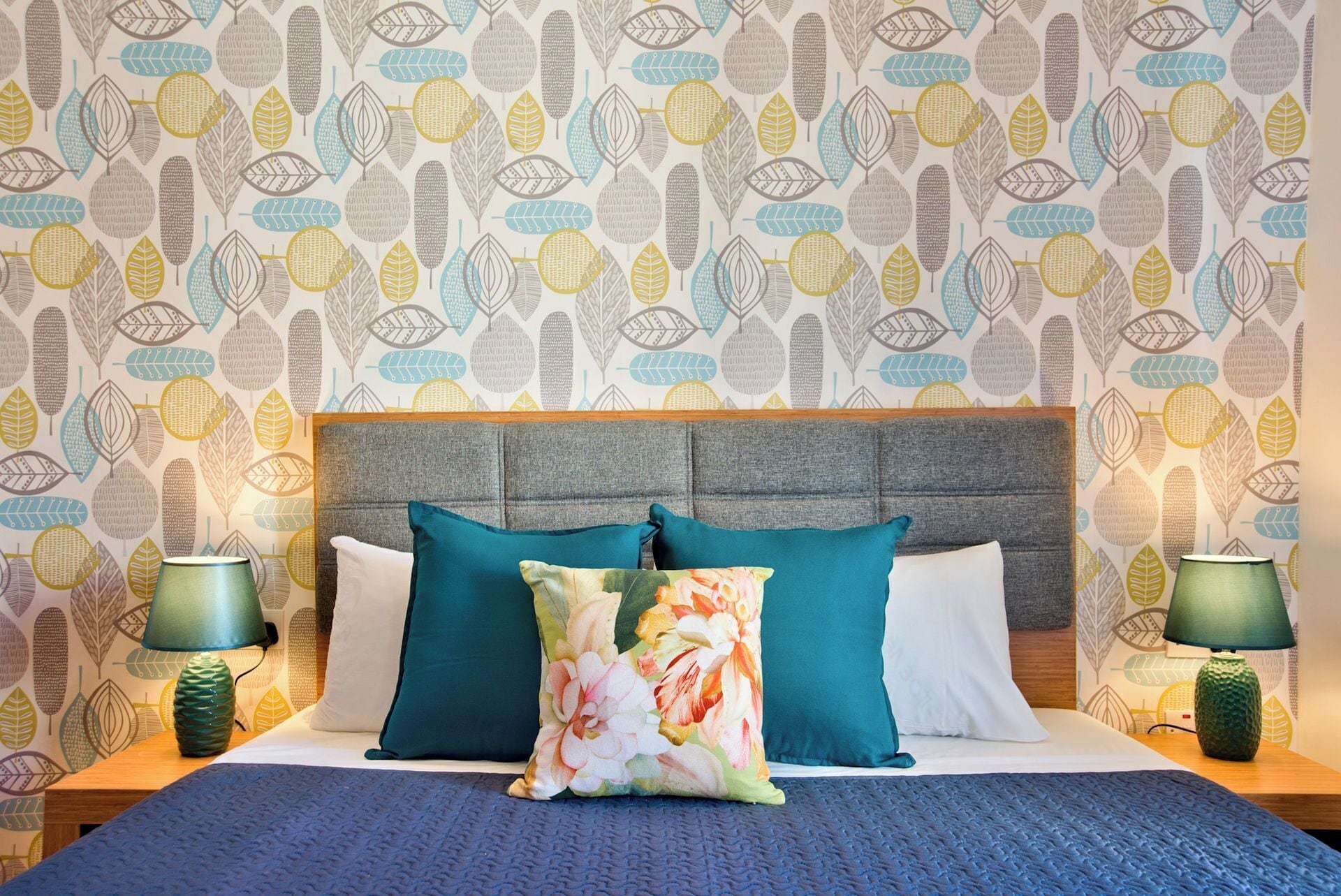 A bedroom with a patterned wallpaper, a grey upholstered headboard, and a bed with teal and white bedding.