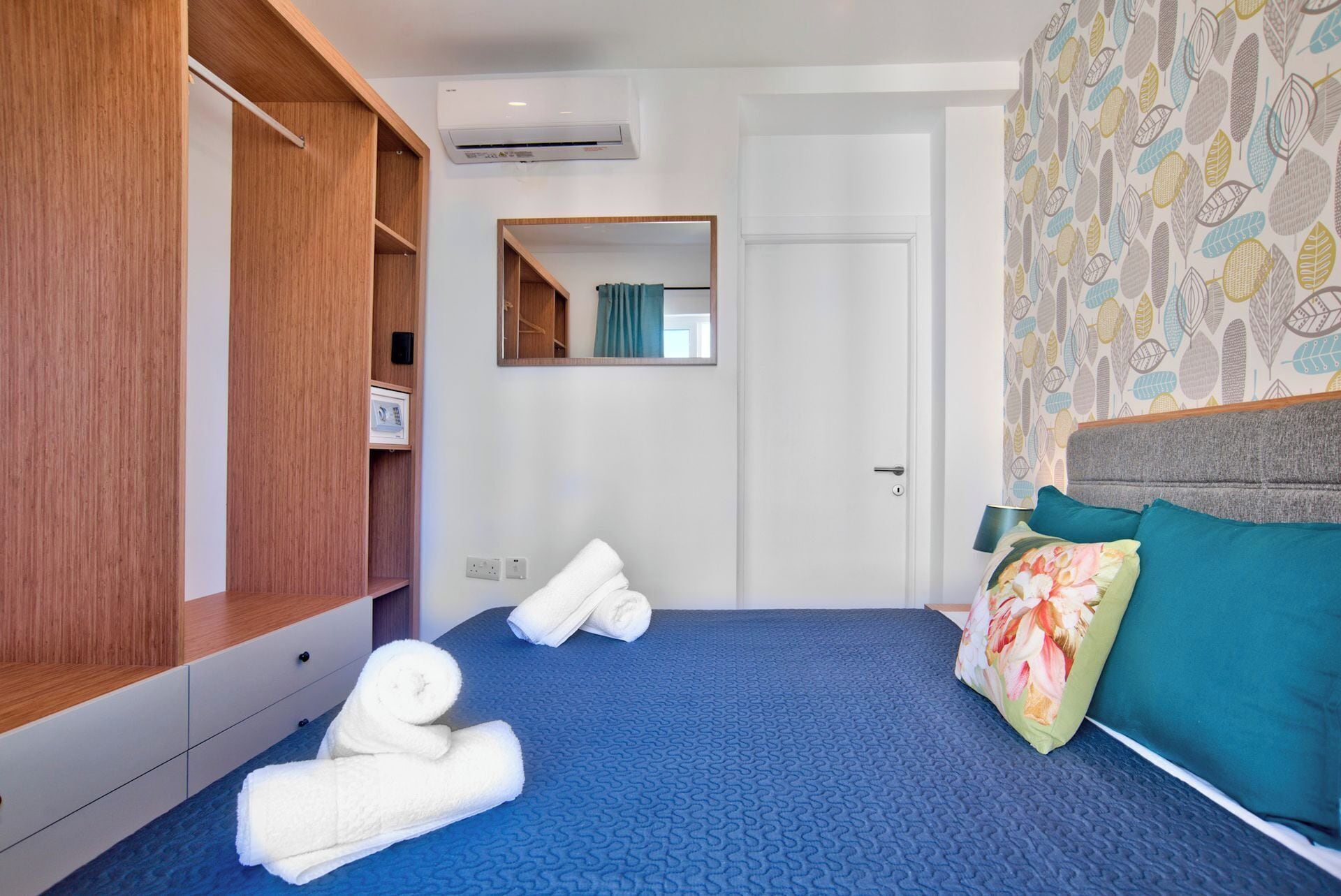 Habitación de hotel con edredón azul texturizado, un armario de madera, un espejo y una puerta.